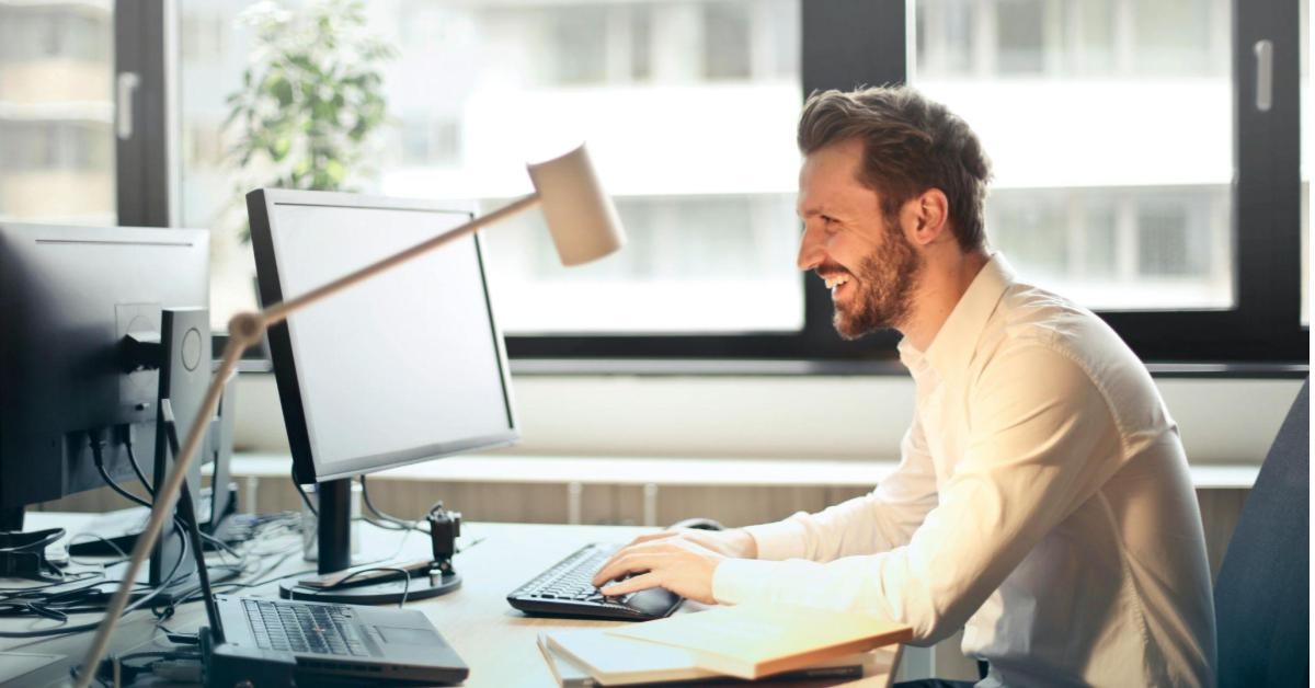 A imagem mostra um homem branco, que veste camisa bege. Ele está em um escritório, sentado de frente para uma grande janela. Na imagem, vemos ele de frente para um monitor de computador, mexendo no mouse. Ele sorri enquanto faz o processo, uma alusão a uma pessoa empreendedora que utiliza integrações para otimizar seu dia a dia.